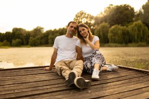 A couple sitting together on a wooden jetty by the lake at sunset, enjoying a warm summer evening.