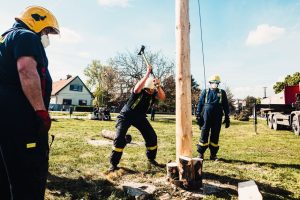 Firefighters using sledgehammers in an outdoor lumberjack demonstration with wooden pole.